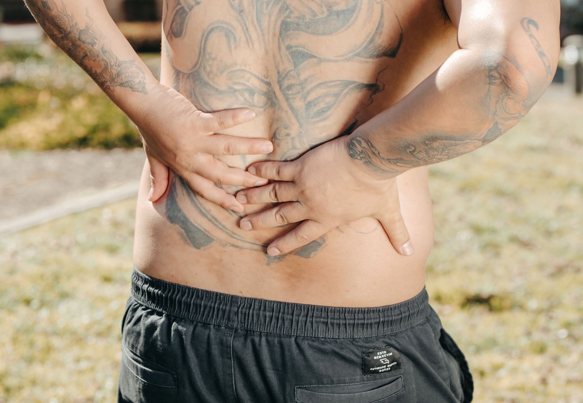 A man with a large tattoo on his back holds his lower back outdoors, showing discomfort.