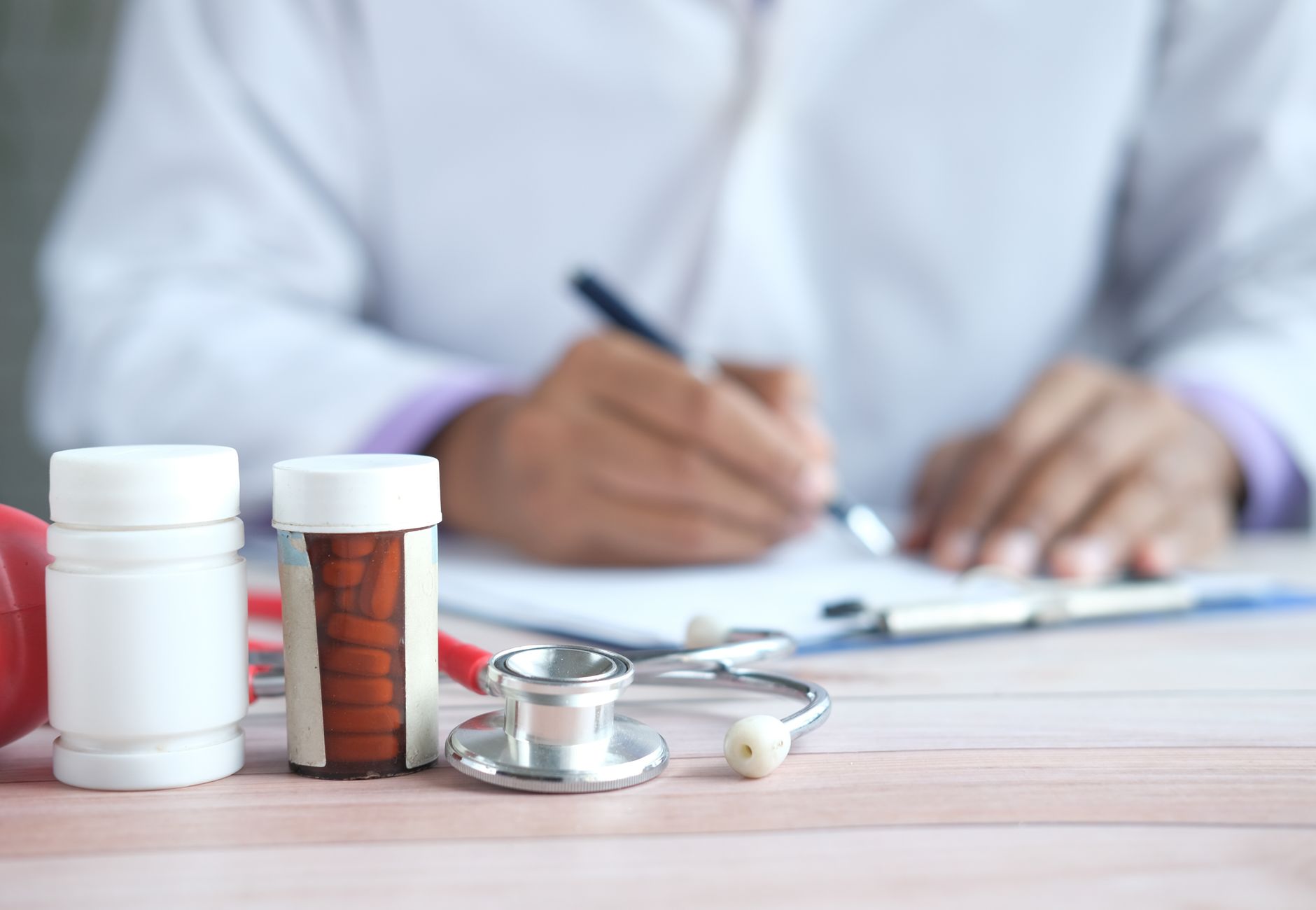 Close-up of doctor writing prescription with medicines and stethoscope on desk.