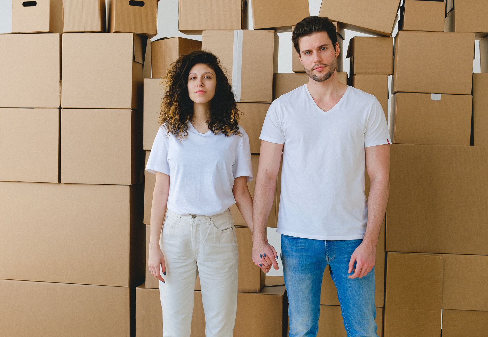 A young couple holding hands in front of a stack of cardboard boxes, possibly indicating moving or relocation.