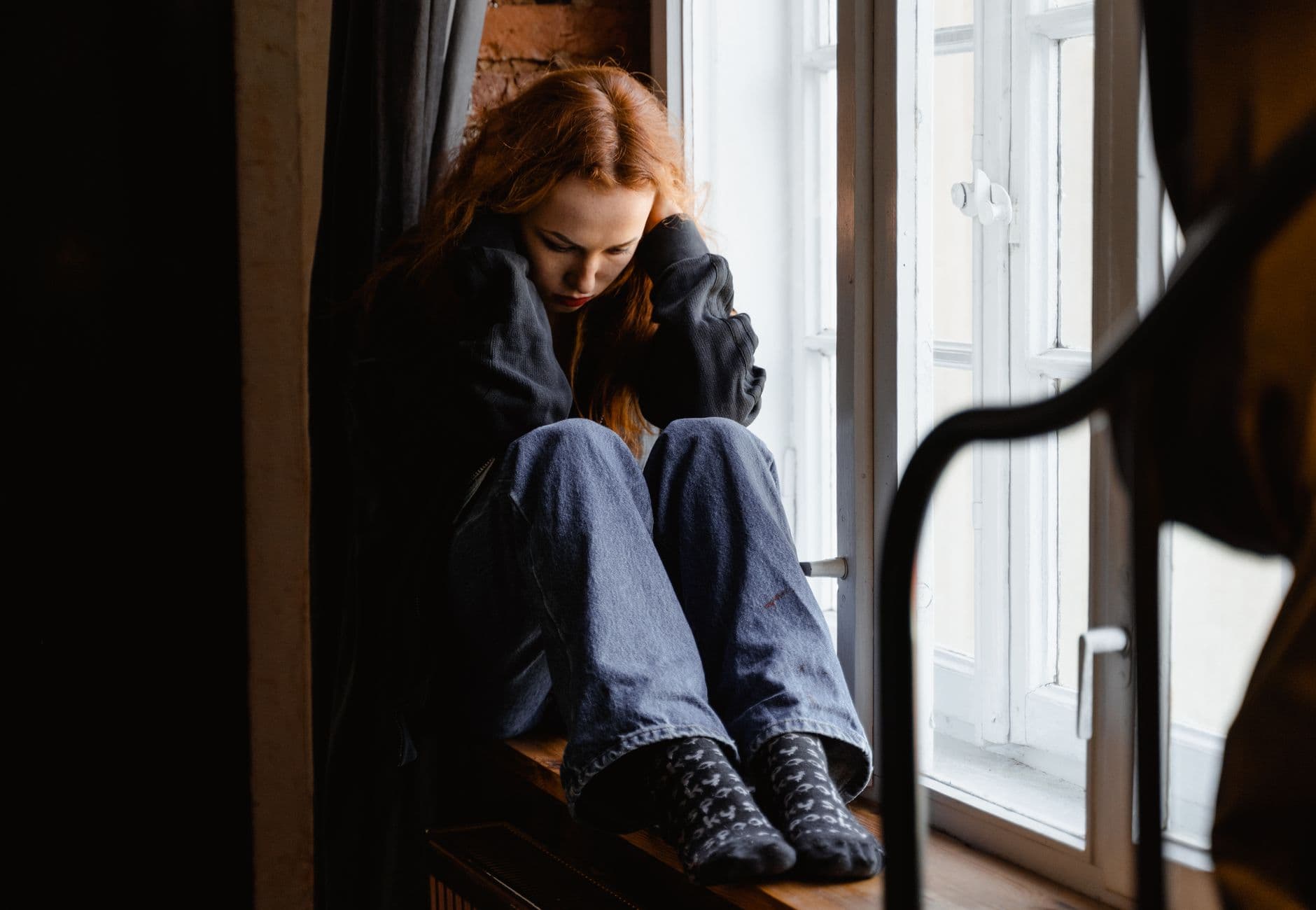 A woman sits pensively by a window, expressing feelings of solitude and contemplation.