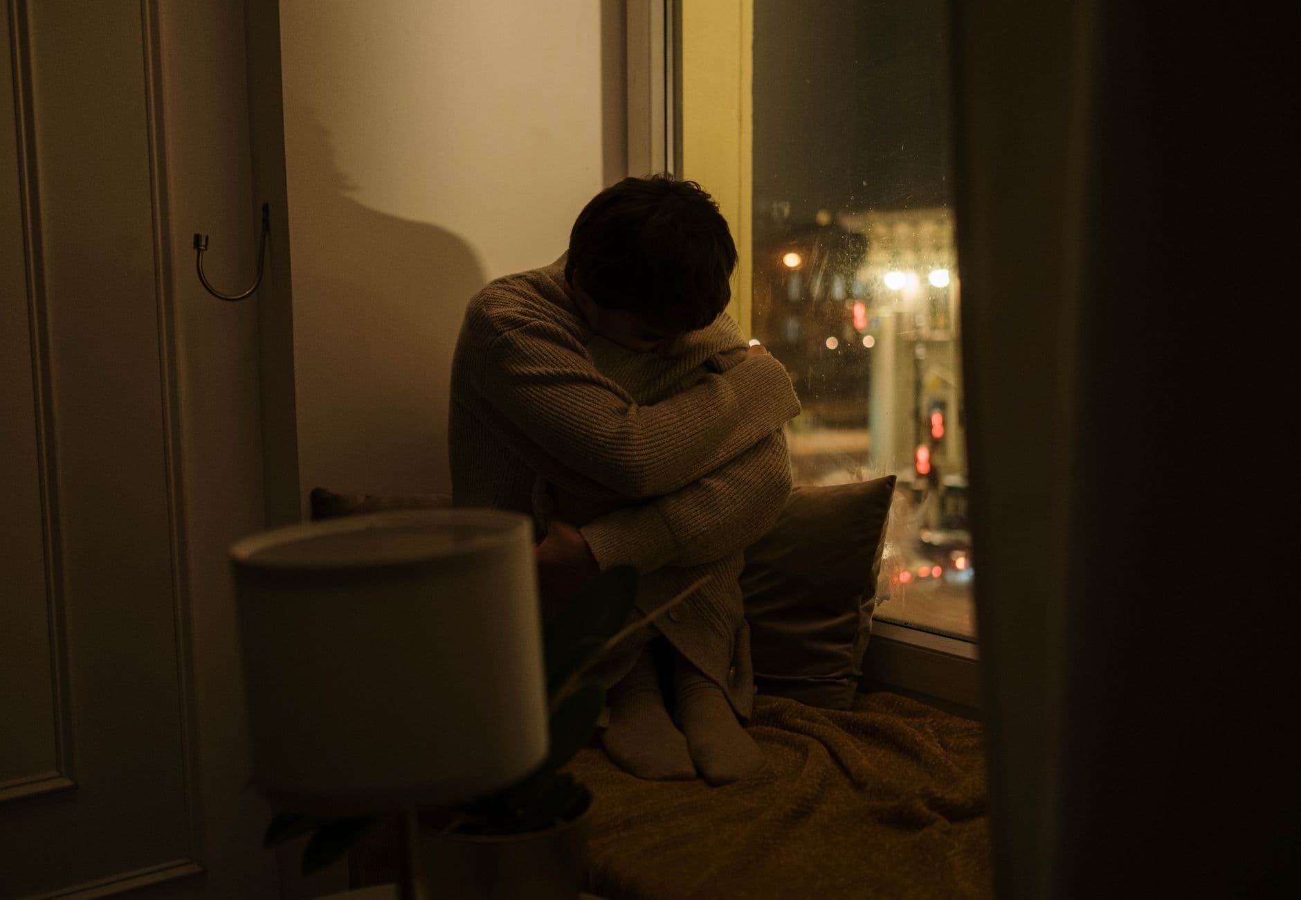 A woman sits alone by a window at night, depicting solitude and reflection.