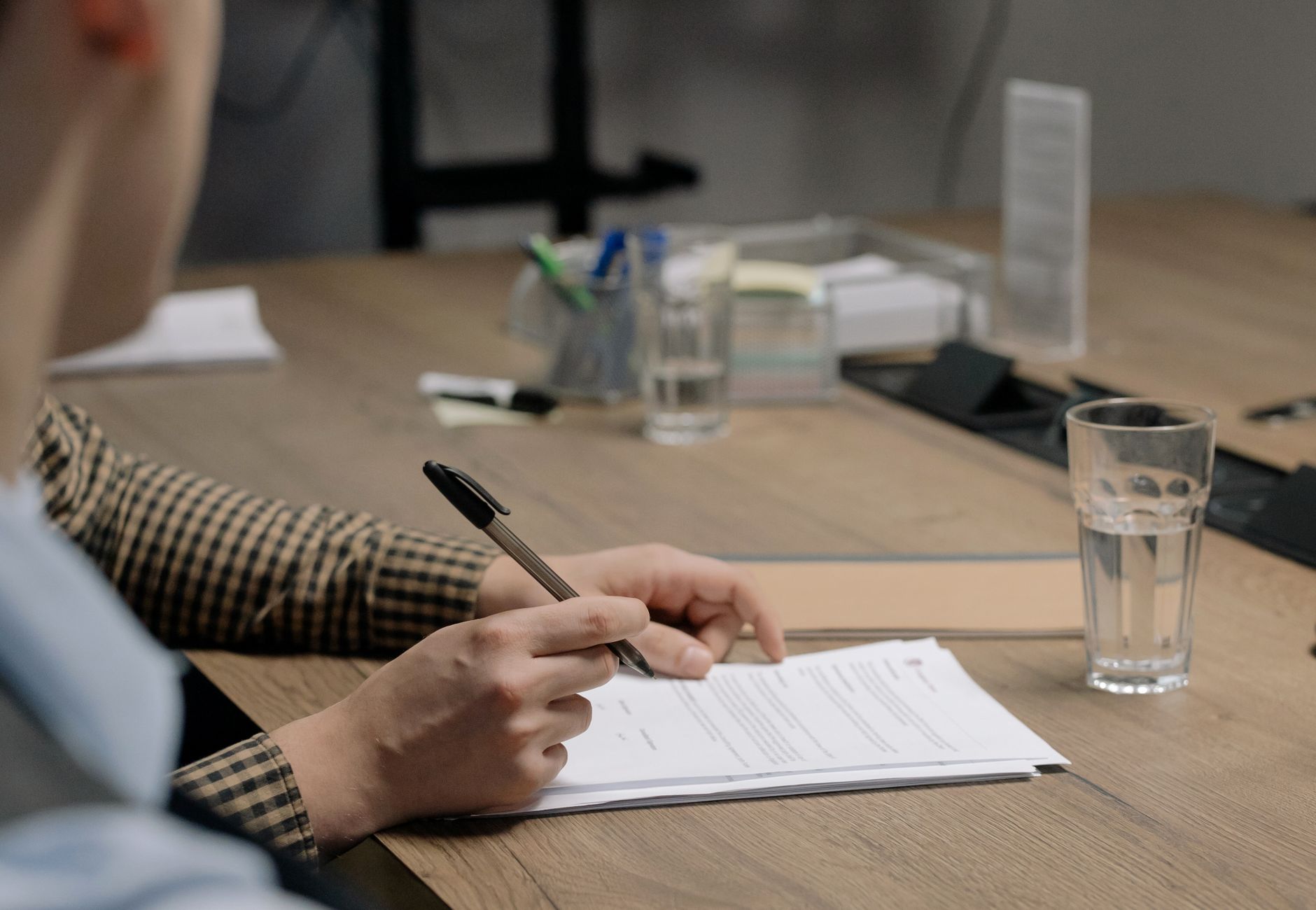 Person in business attire signing a document at a wooden table in an office setting.
