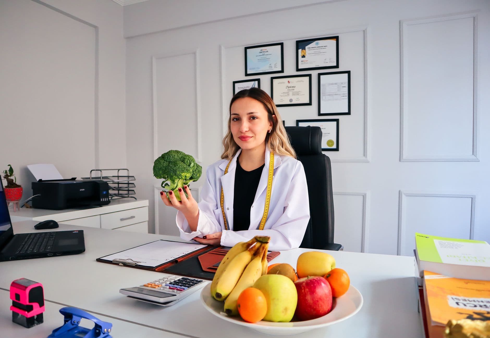 Female nutritionist in office holding broccoli, surrounded by fruits, promoting healthy lifestyle.