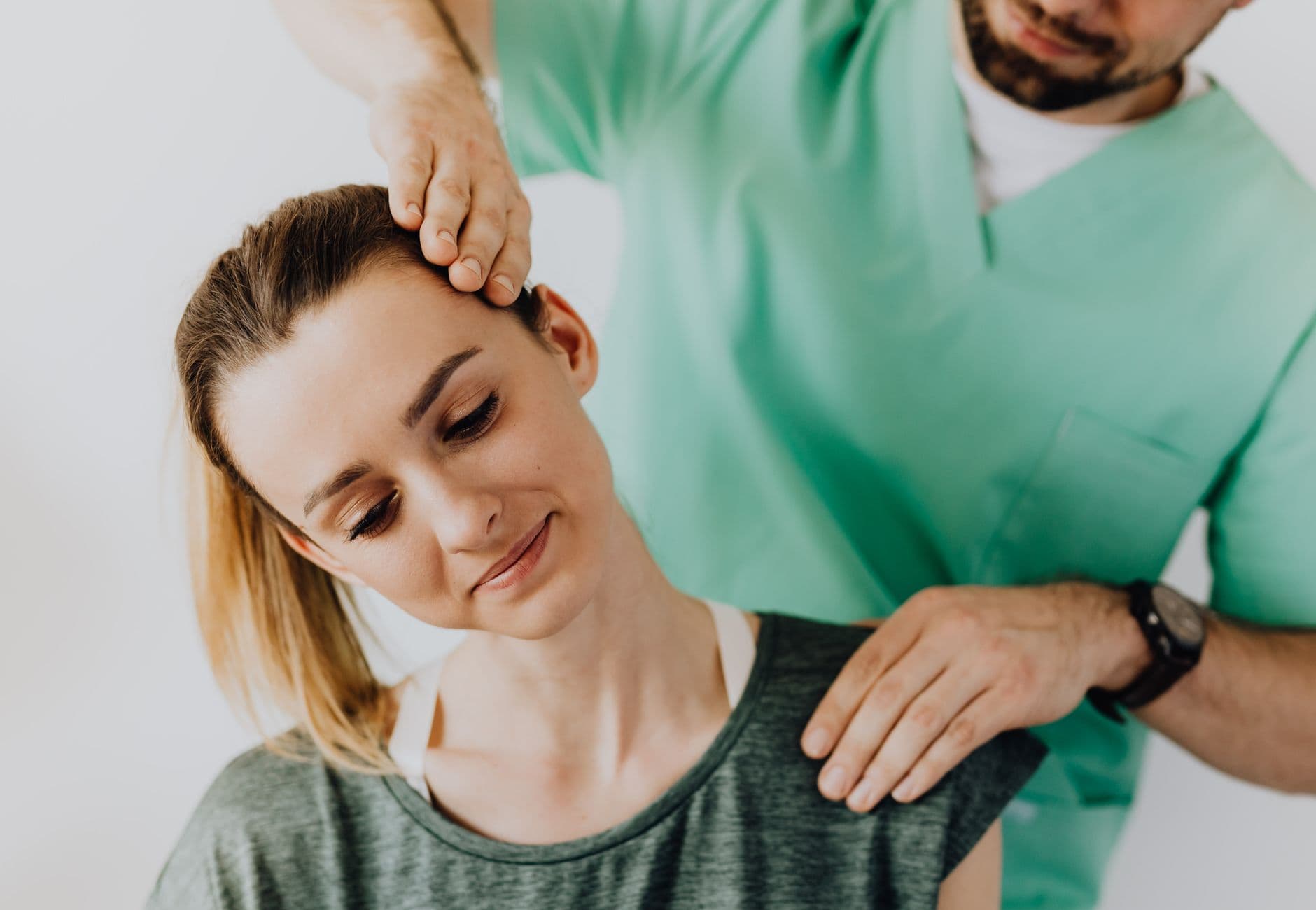Serious doctor in uniform massaging smiling female patients neck and stretching stiff neck muscles against gra