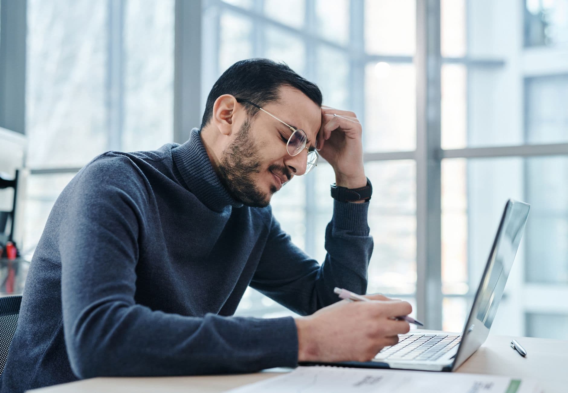 Man looking stressed at laptop in a modern office with large windows.